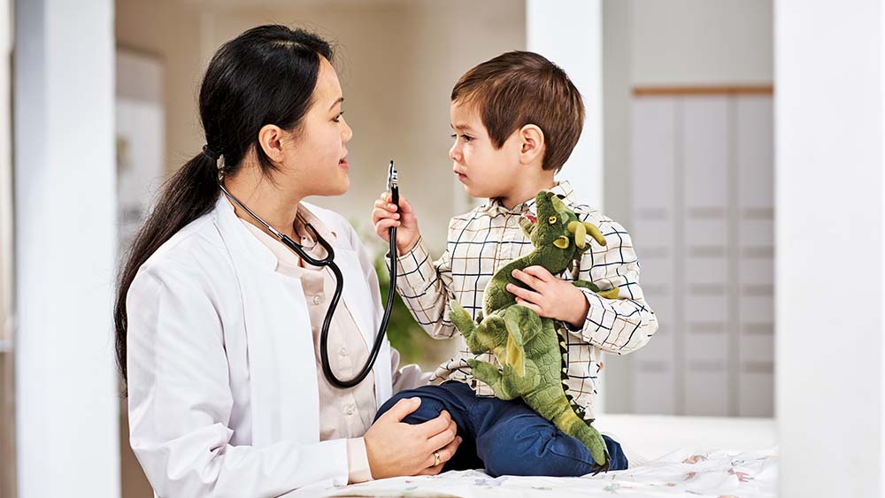 Una profesional sanitaria habla con un niño pequeño que sostiene un dinosaurio de juguete. El niño parece comprometido y curioso, mientras que el profesional sanitario tiene una expresión cálida y amistosa