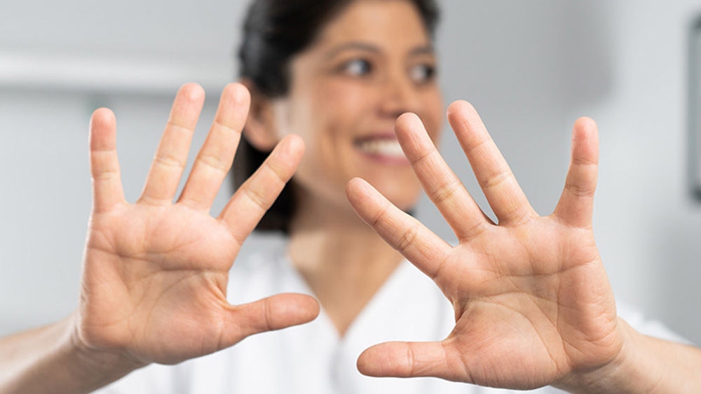 Una persona lleva una bata de laboratorio blanca y mira a la cámara. Las dos manos se extienden hacia delante con los dedos separados. La cara de la persona se desfoca intencionadamente para proteger su identidad. En segundo plano, hay un entorno interior borroso con una imagen colgada en la pared y un mueble que parece una cama o un sofá.