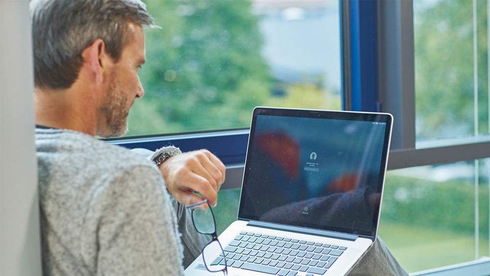 Una persona sentada sostiene un ordenador portátil en el regazo. Con la mano izquierda, escriben en el teclado mientras sostienen un par de gafas en la mano derecha. En la pantalla del portátil, se ve un símbolo borroso que se asemeja a unos auriculares, junto con la etiqueta «Tap to listen» (Pulse para escuchar). En segundo plano, se puede ver un paisaje verde y natural a través de una gran ventana.