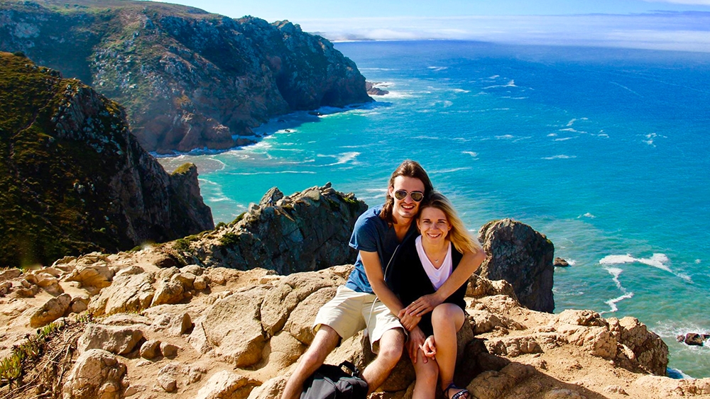 Un hombre y una mujer se sientan juntos en una roca, sonriendo, con el mar al fondo y el pelo soplando al viento
