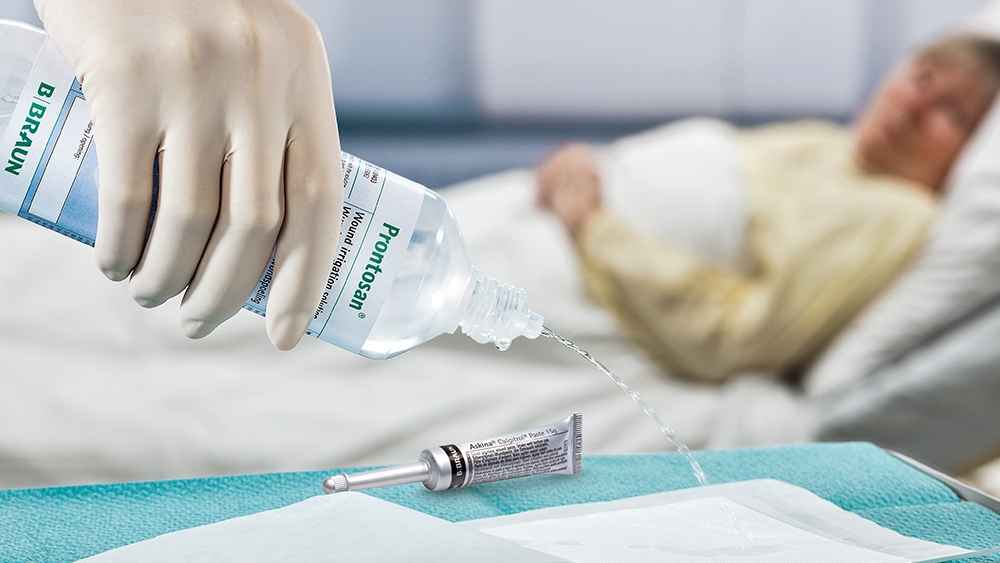 En primer plano, una mano con guante médico vierte Prontosan Irrigation Solution sobre una almohadilla que se encuentra sobre la mesa junto a un tubo de Askina Calgitrol. En segundo plano, una paciente está tumbada en una cama de hospital.