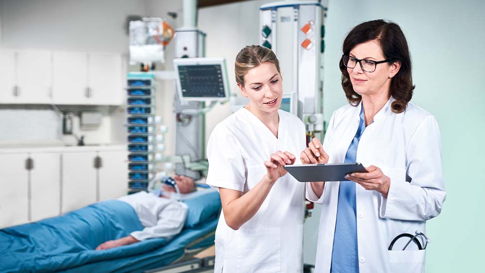 Un médico y una enfermera discuten la dosis de la infusión inteligente de B. Braun, que se introduce directamente desde una tableta. El paciente de la UCI se puede ver en segundo plano, conectado a una torre llena de dispositivos de infusión inteligentes.