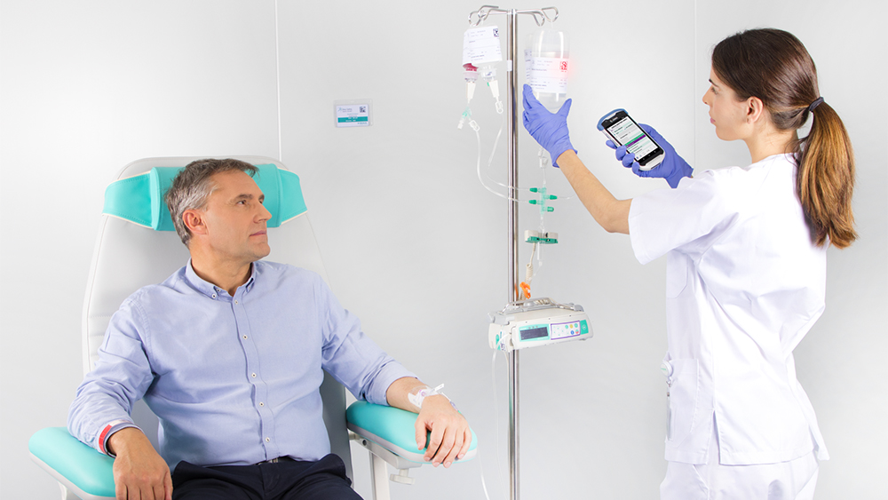 La imagen muestra a un paciente sentado en una silla recibiendo una infusión de un dispositivo médico de B. Braun, mientras se ve a una enfermera configurando la dosis de infusión en el dispositivo.