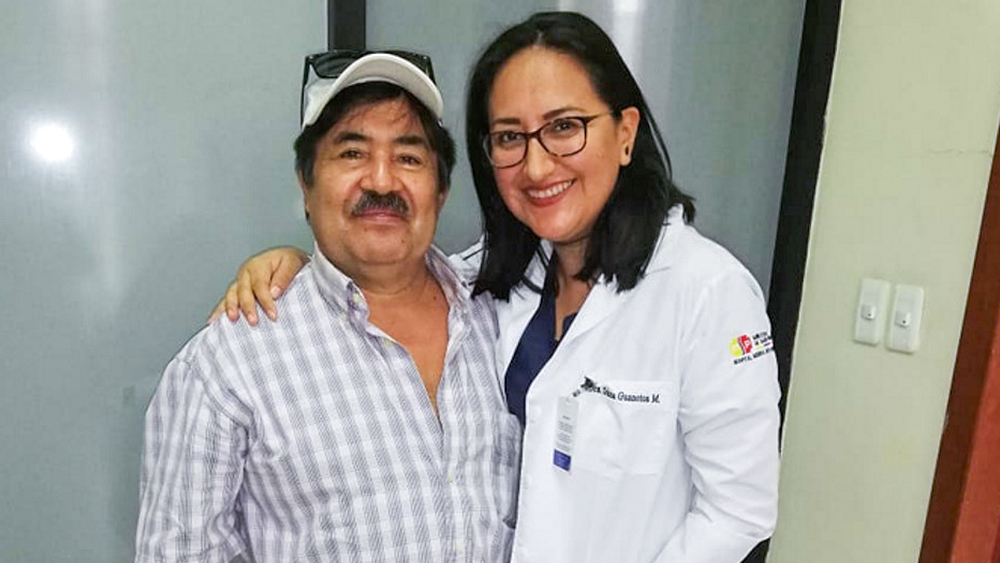 Un hombre y una mujer están uno al lado del otro sonriendo. La mujer sujeta el brazo alrededor del hombre