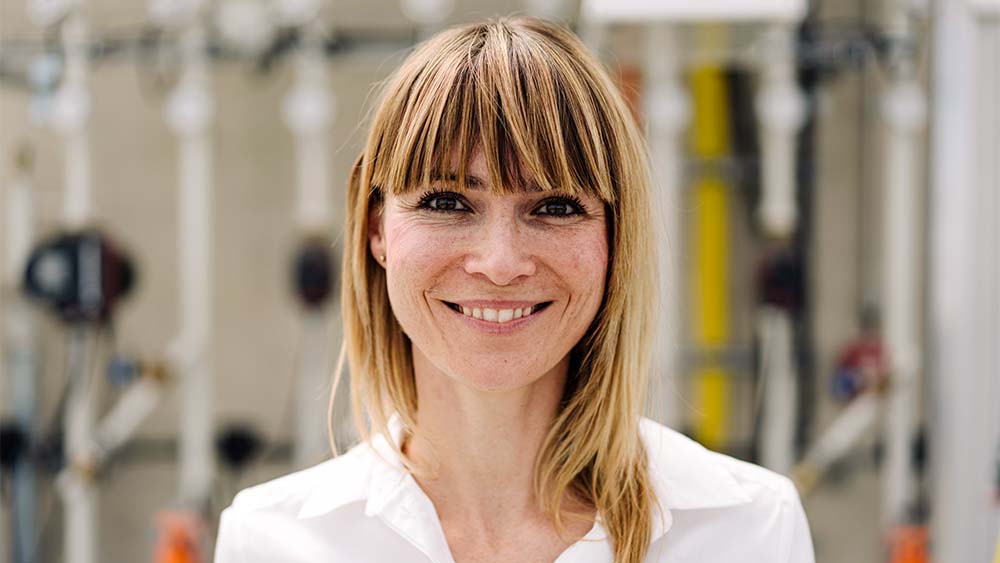 Primer plano de una mujer mirando a la cámara, sonriendo y llevando una camisa blanca en un entorno brillante. 