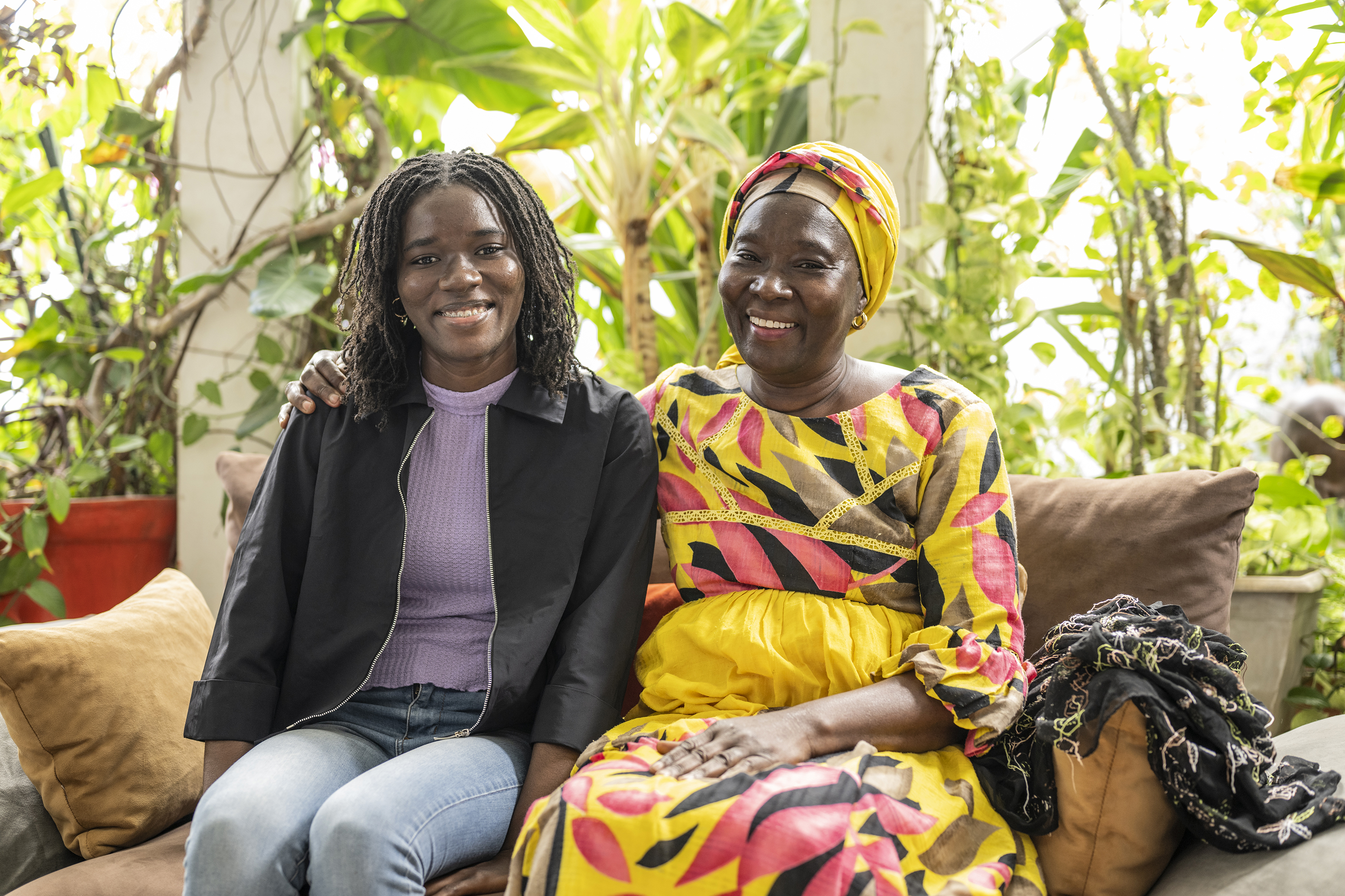 Silberia Diack y su madre están sentadas en un sofá en una habitación bien iluminada. Se sujetan entre sí en los brazos y sonríen mirando hacia delante. 